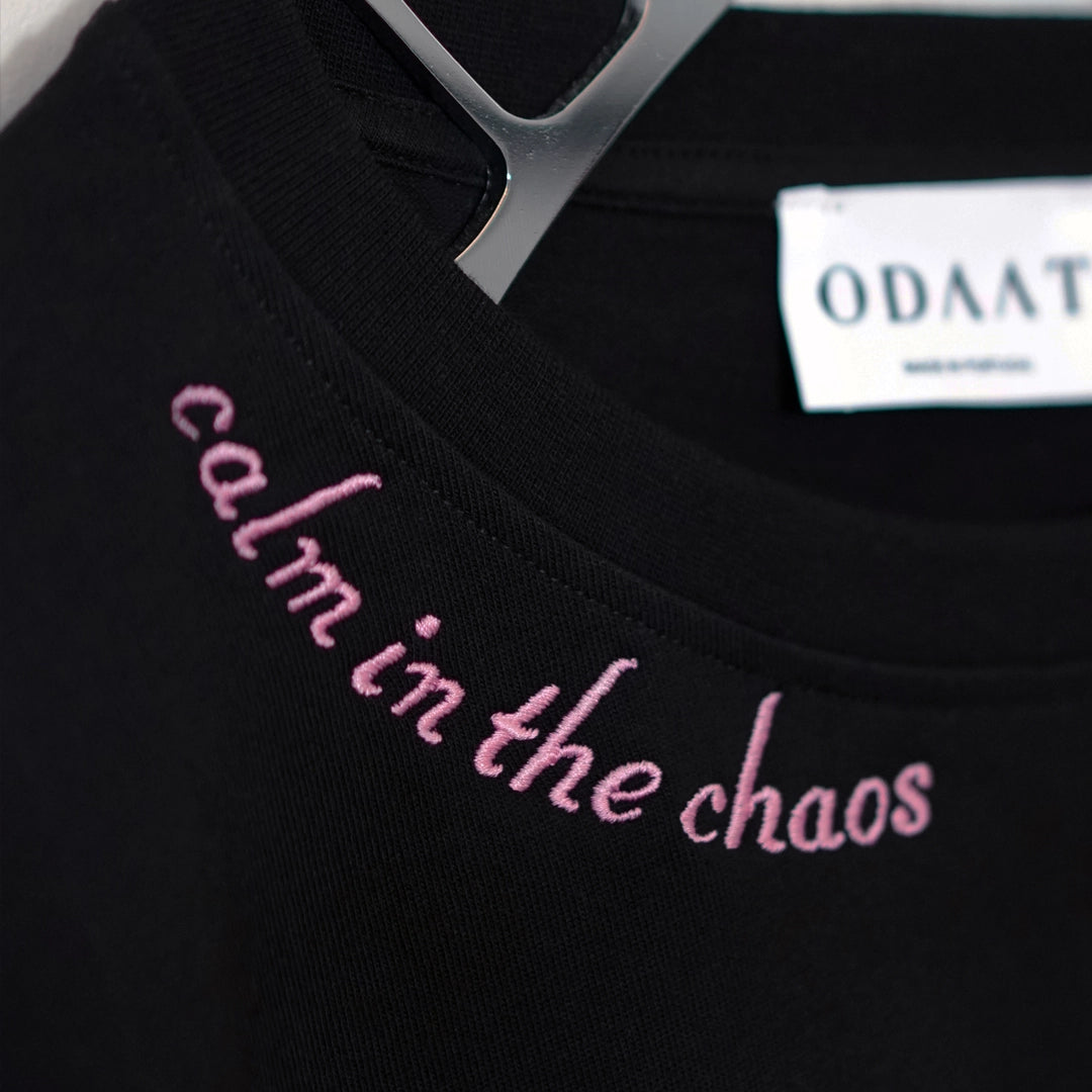 Close-up of the Calm in the Chaos Tee in black, featuring soft pink embroidered ‘calm in the chaos’ text along the neckline and an ODAAT inner label.