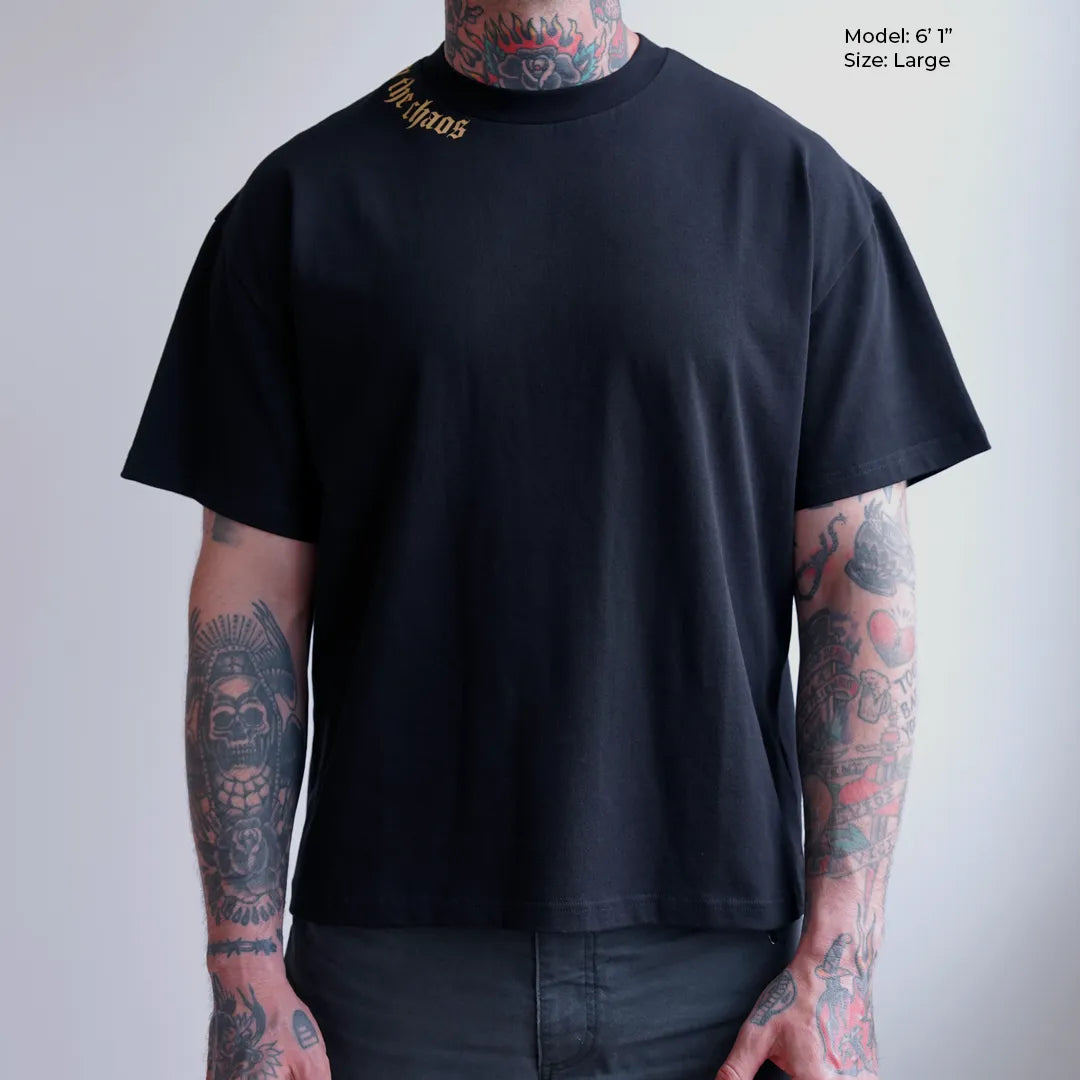 Person wearing black calm in the chaos t-shirt with visible tattoos on arms against a plain background