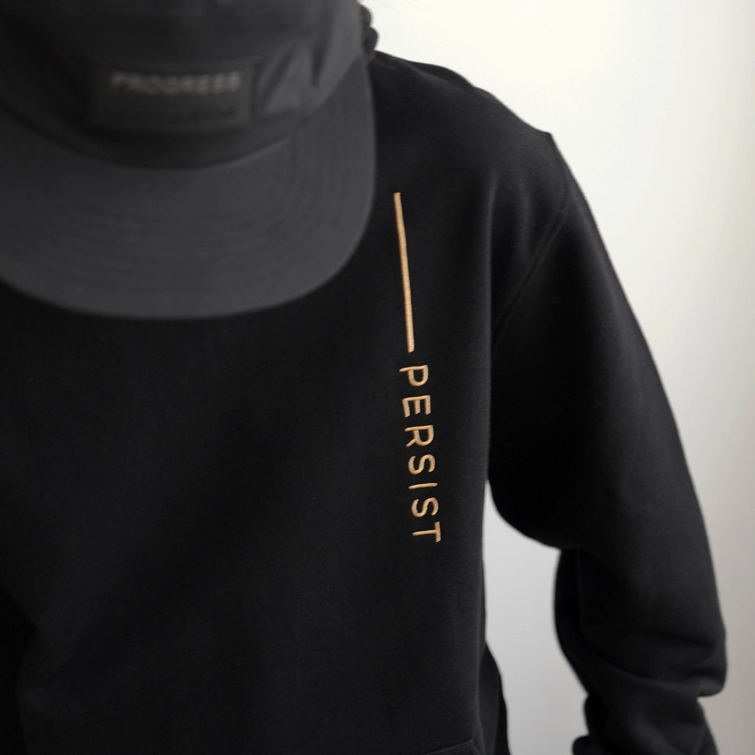 ODAAT Apparel black hoodie with vertical gold lettering that reads “PERSIST” on the chest. Minimalist streetwear design styled with a matching black cap, emphasizing resilience and daily discipline.