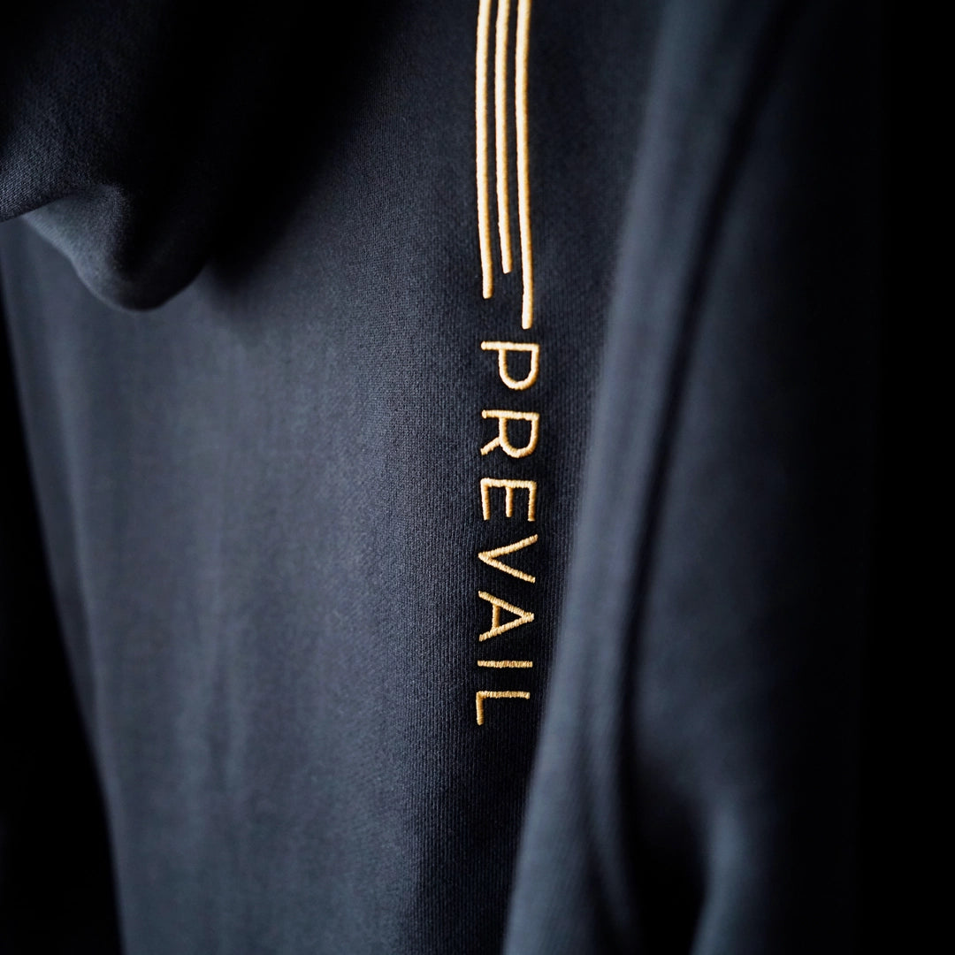 Close-up of the Persist.Prevail Hoodie featuring vertical gold ‘PREVAIL’ embroidery on black fabric—a subtle tribute to quiet resilience.