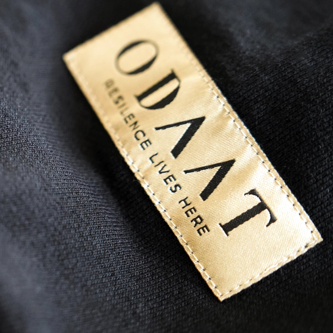 Close-up of ODAAT’s gold ochre woven sleeve label reading ‘Resilience Lives Here,’ stitched onto black fabric as a symbol of daily perseverance.