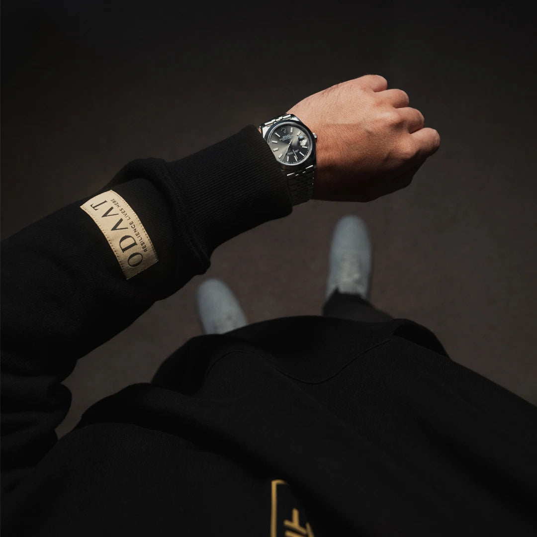 Close-up of ODAAT’s gold ochre woven sleeve label reading ‘Resilience Lives Here,’ stitched onto black fabric as a symbol of daily perseverance.