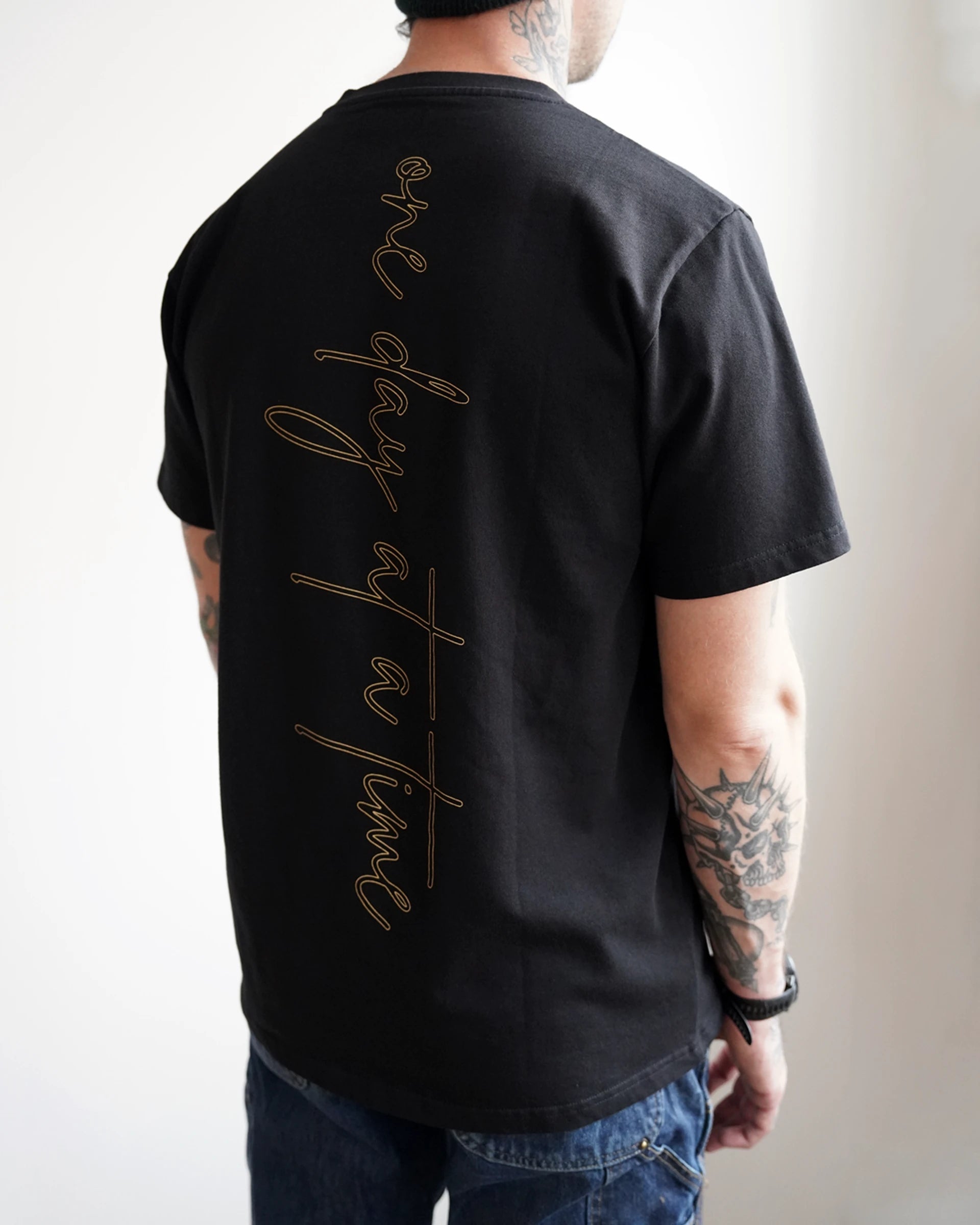 Model wearing the ODAAT “One Day At A Time” Script Tee in black, showcasing the vertical gold script design down the back — minimal, elevated, and built on resilience.