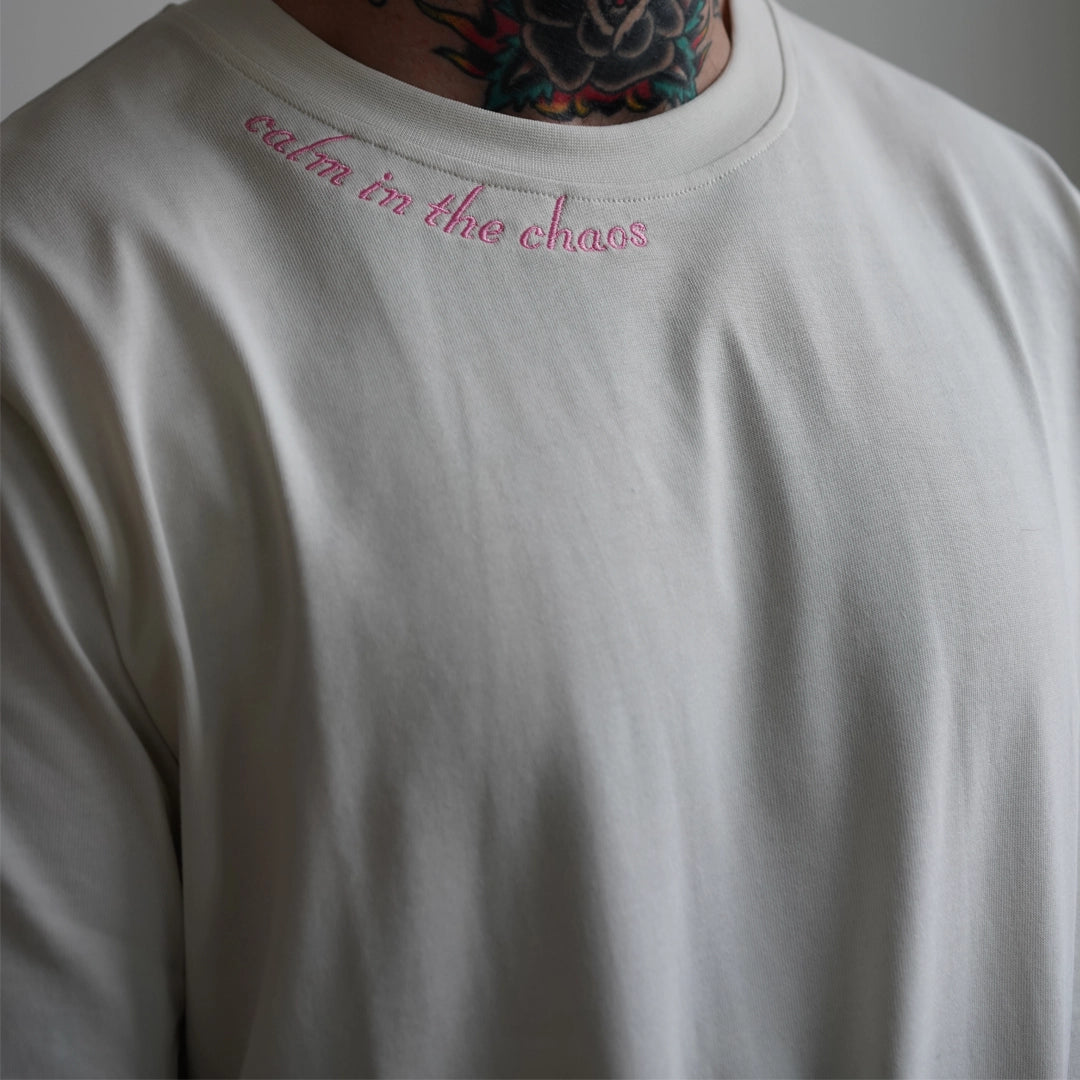 Close-up of the Calm in the Chaos Tee in bone white, featuring soft pink embroidered text at the collar reading ‘calm in the chaos’—crafted from breathable organic cotton.
