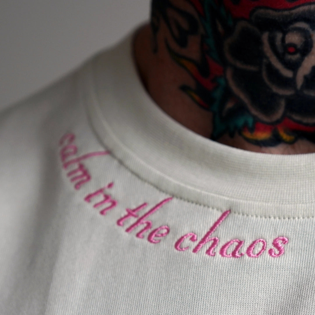 Detailed close-up of the Calm in the Chaos Tee’s soft pink embroidered neckline text, stitched on bone white organic cotton as a quiet statement of inner strength.