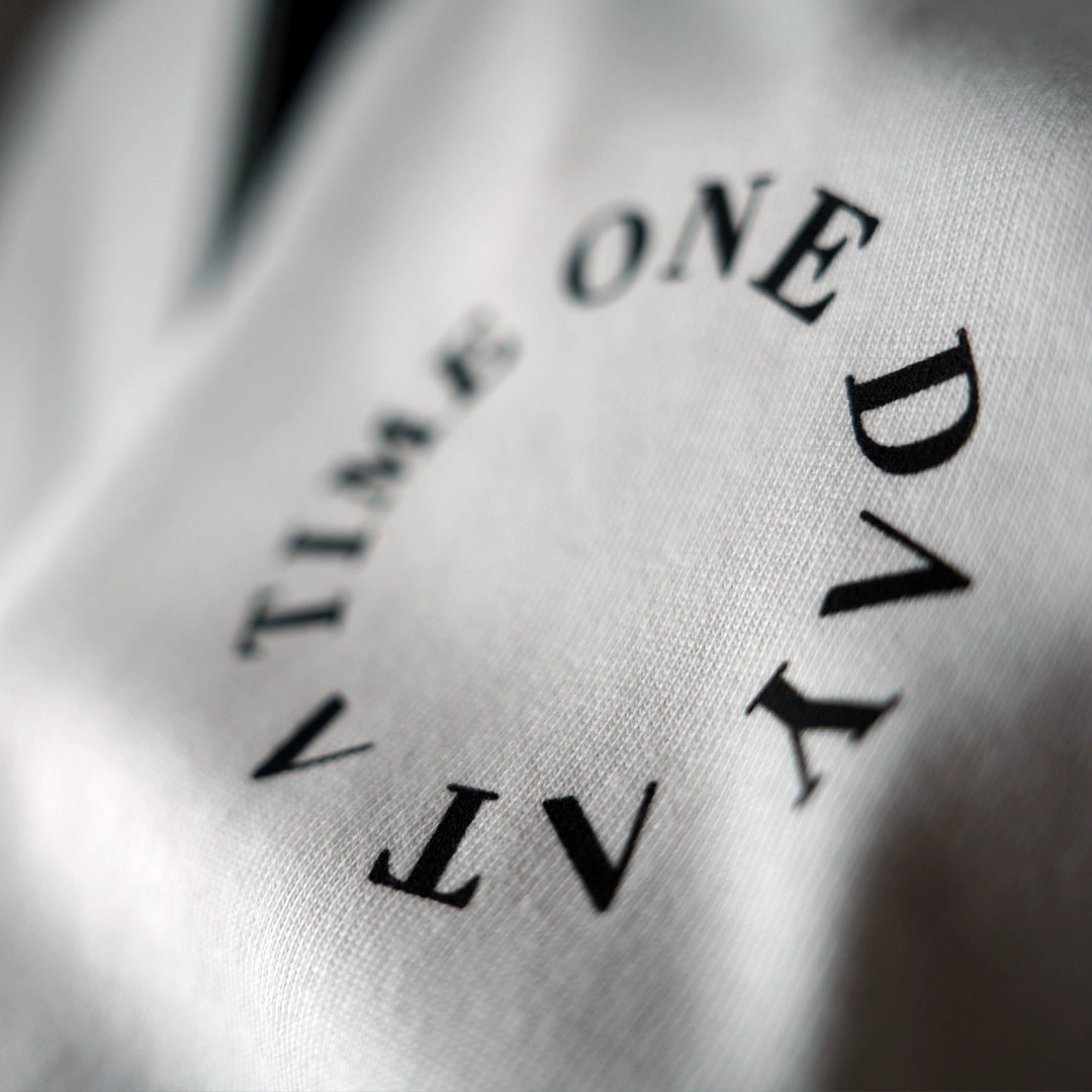 Close-up of white ODAAT Apparel t-shirt with black circular ‘One Day At A Time’ text on chest — minimalist design symbolizing resilience, sobriety, and intentional living in premium streetwear style.