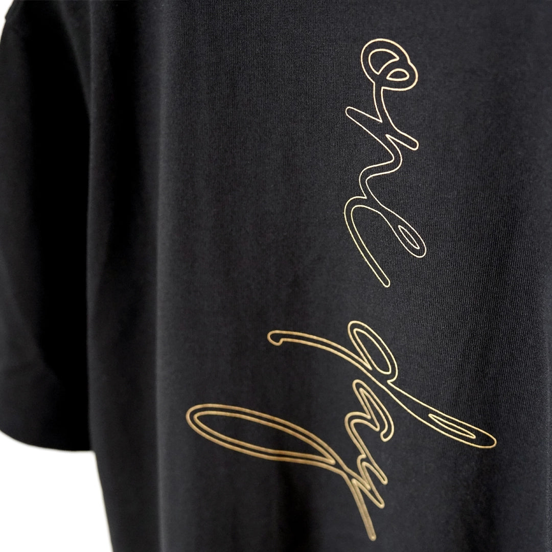 Close-up of the ODAAT Script Tee featuring vertical ‘one day’ lettering in gold ochre script on soft black organic cotton.