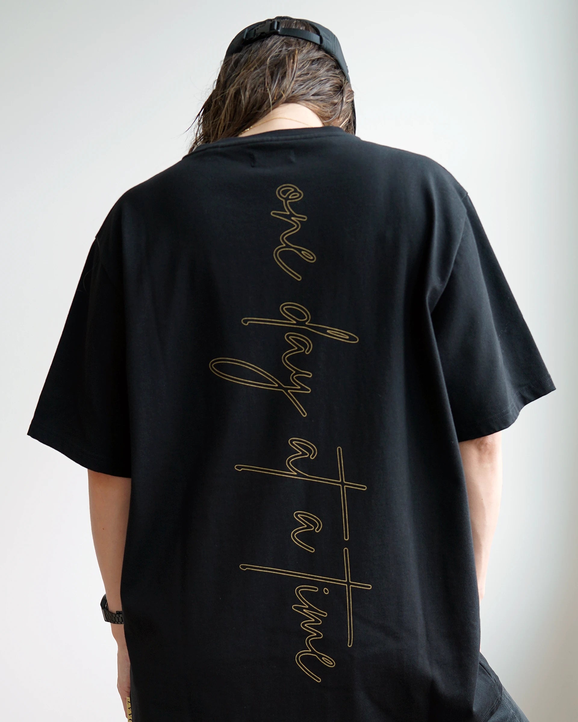 Model wearing the ODAAT “One Day At A Time” Script Tee in black, featuring oversized vertical gold script lettering down the back, representing resilience and mindful minimalism.