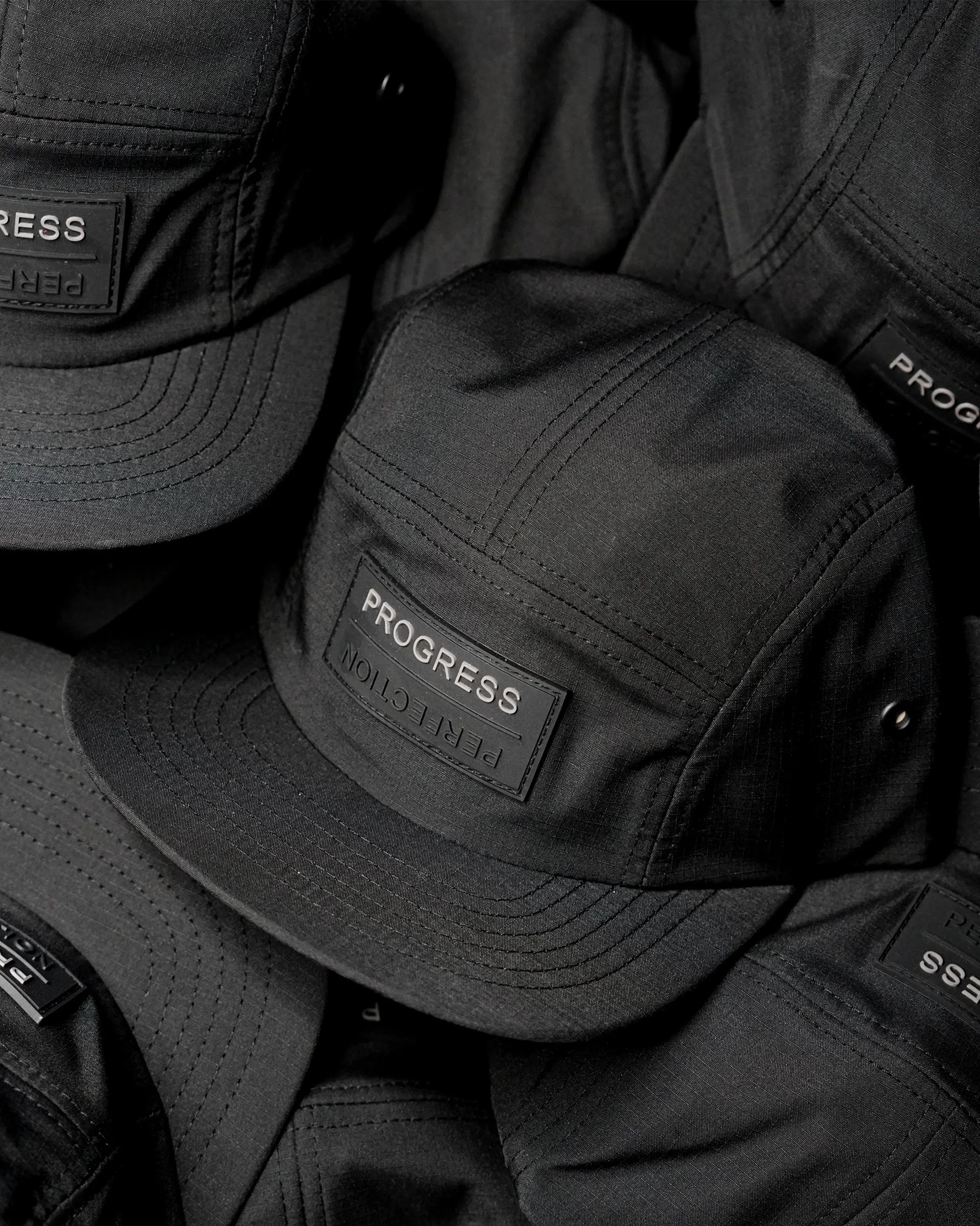 Close-up of stacked black performance caps with PROGRESS branding, highlighting material texture, stitching, and structured construction