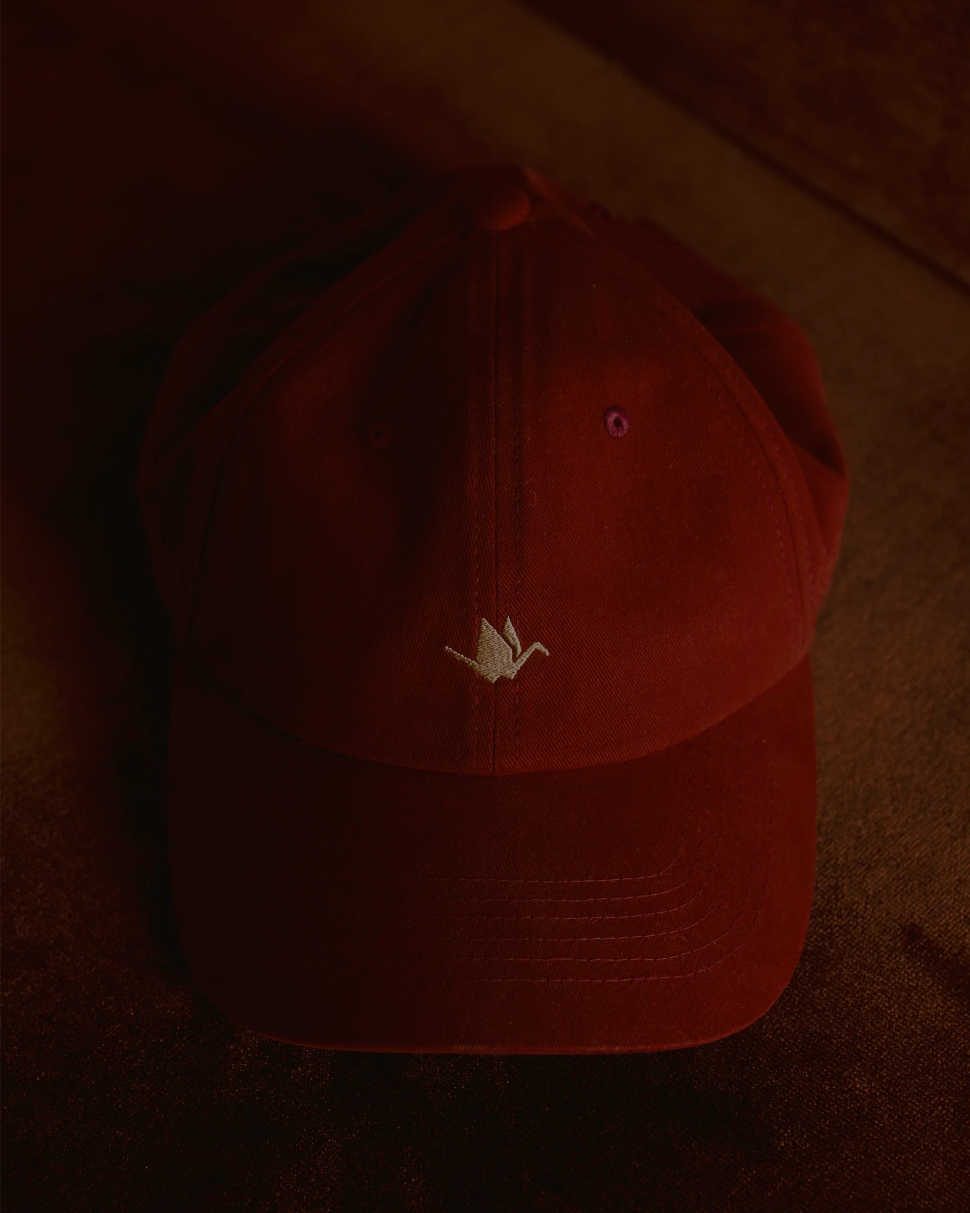burgundy signature dad cap close-up showing curved brim and panel construction, soft cotton fabric with crane embroidery, minimal product detail shot with warm low light background