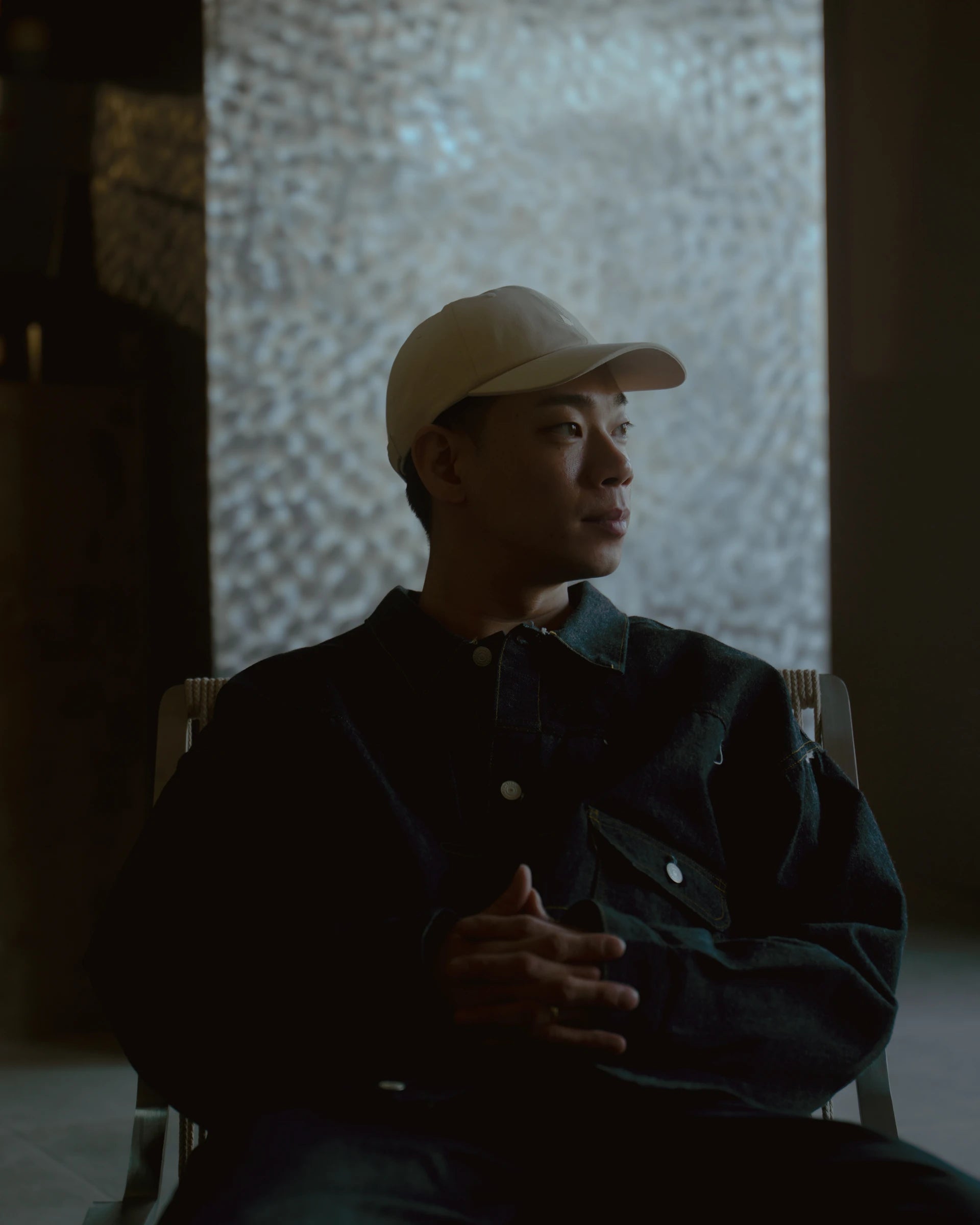 rice paper signature dad cap worn by model, seated portrait with side gaze, soft cotton cap with curved brim, neutral tone contrasted against dark jacket in dim interior with textured wall backdrop