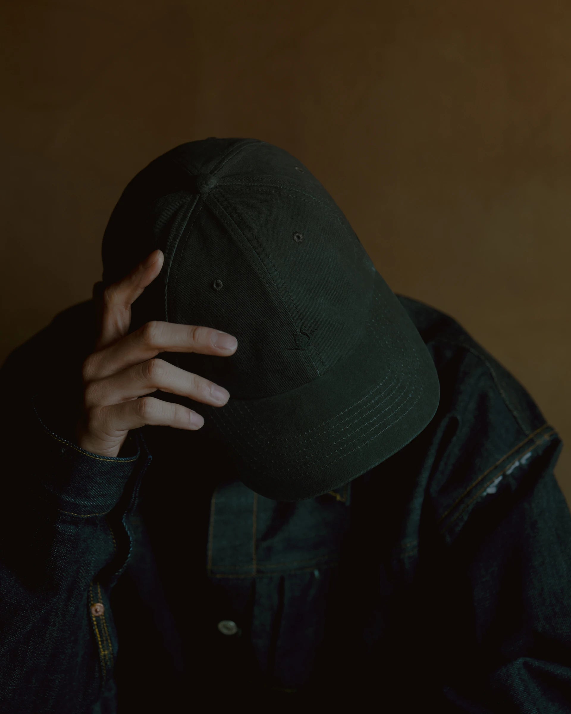 washed black signature dad cap worn low with hand adjusting brim, soft structured cotton hat with curved visor, minimal crane embroidery, moody lifestyle portrait with warm tones and shadowed face