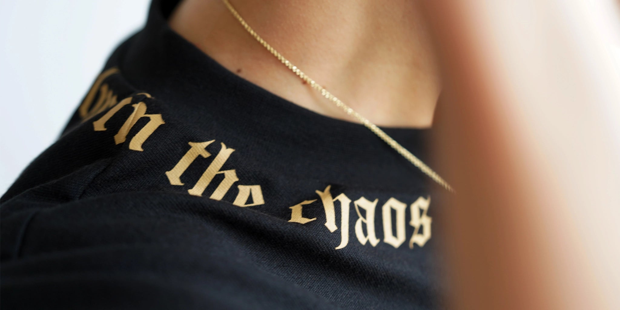 Close-up of black boxy tee with gold “Calm in the Chaos” neckline print, worn with a thin gold chain.