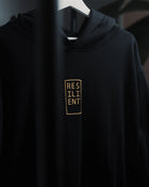 Minimal black Resilient Hoodie by ODAAT Apparel, heavyweight organic cotton with gold print logo, hanging on white hanger, premium streetwear design,