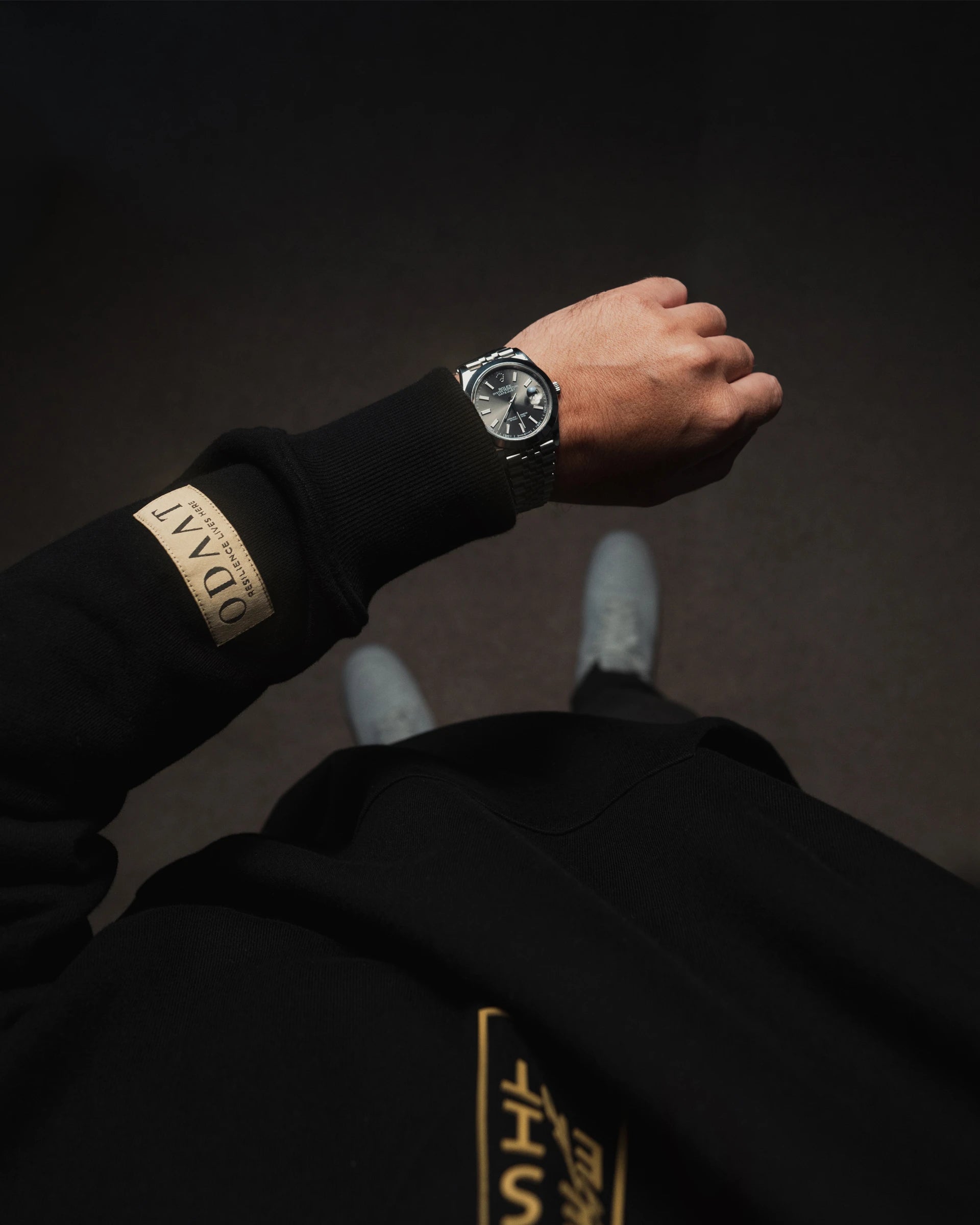 Close-up of ODAAT Apparel Resilient Hoodie sleeve with woven label, black organic cotton streetwear featuring gold details, worn with silver wristwatch.