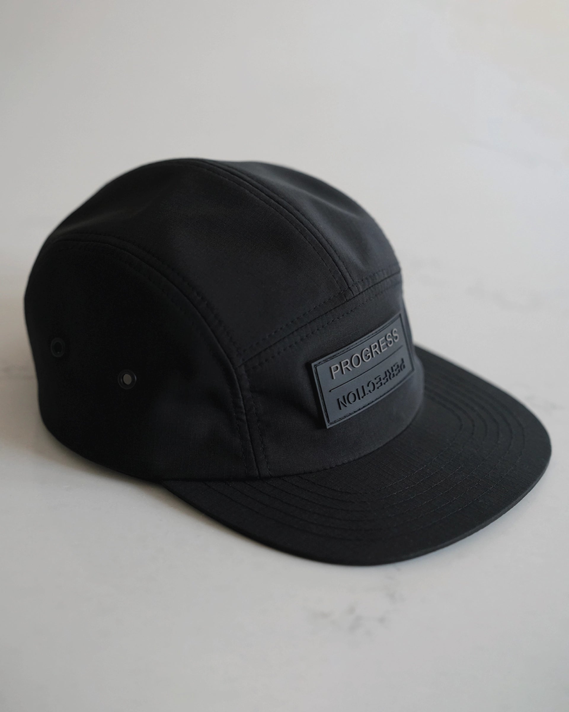 Minimal black 5-panel performance hat with matte rubber ‘Progress Over Perfection’ patch, structured front panel, flat brim, and ventilation eyelets—modern athletic cap by ODAAT Apparel.