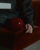 burgundy signature dad cap resting on upholstered chair, soft cotton hat with curved brim and crane embroidery, lifestyle product shot with warm tones and textured fabric background