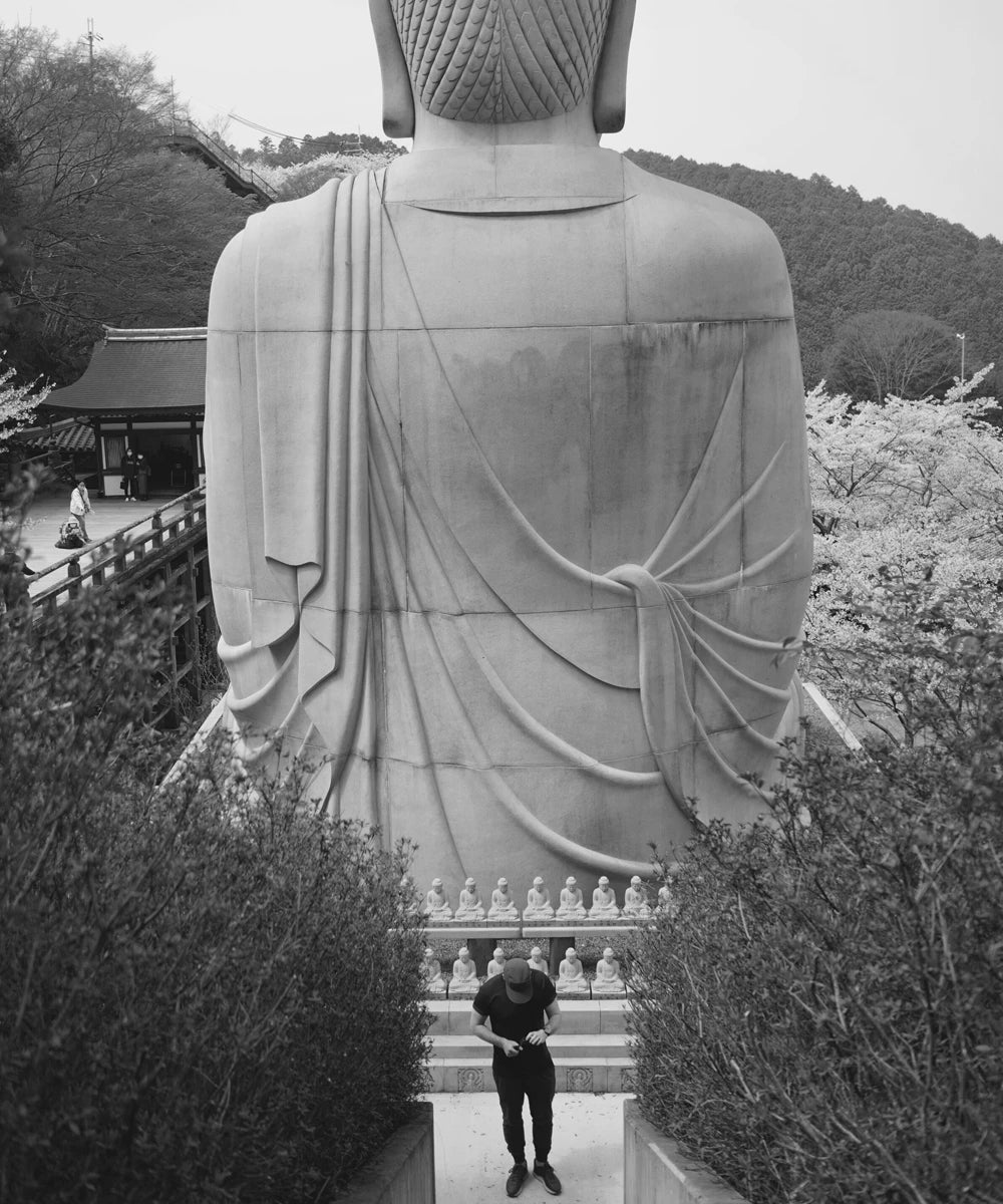 ODAAT founder Jeremy Webster standing beneath a stone Buddha in Kyoto, Japan — black and white founder portrait.