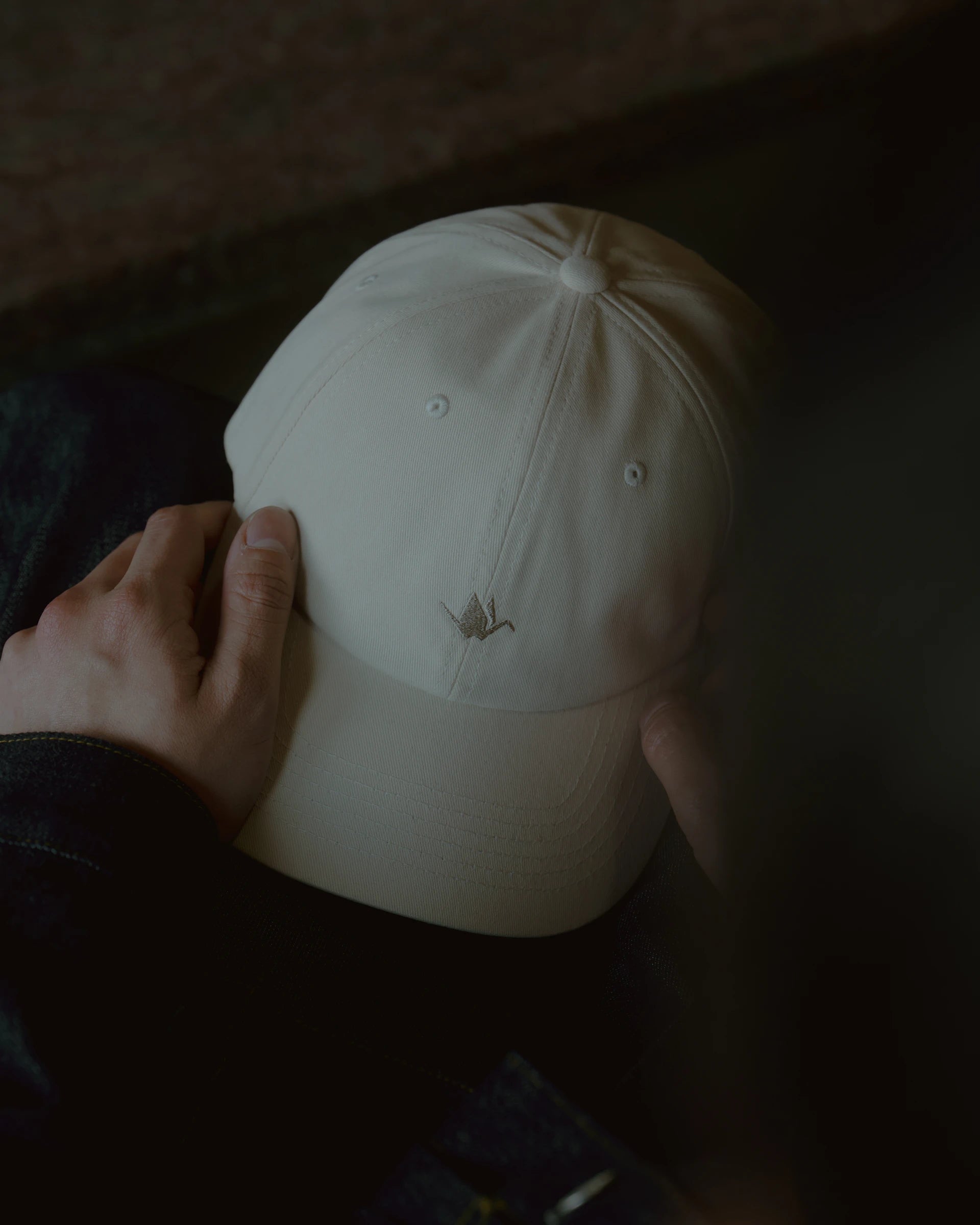 rice paper signature dad cap top view held by hands, curved brim and panel stitching visible with subtle crane embroidery, soft cotton texture in low light against dark background