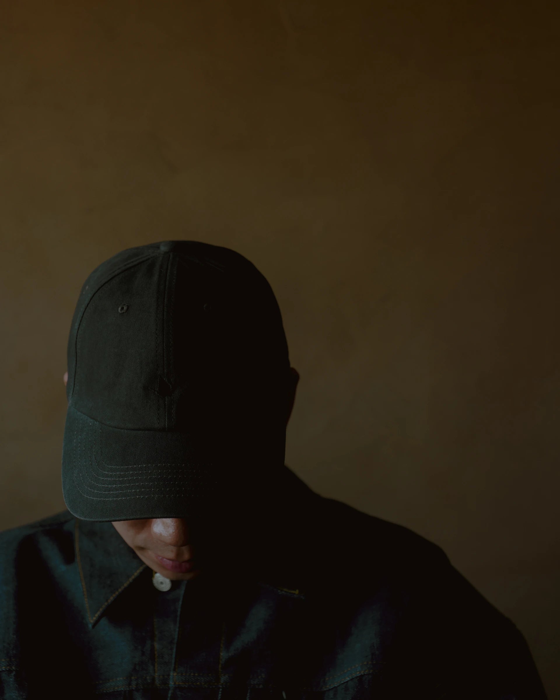 washed black signature dad cap worn low over face, curved brim cotton hat with minimal crane embroidery, soft structured fit, moody portrait with warm backdrop and shadowed lighting