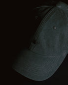washed black signature dad cap close-up showing curved brim and panel construction, soft cotton fabric with subtle crane embroidery, minimal product detail shot on dark background