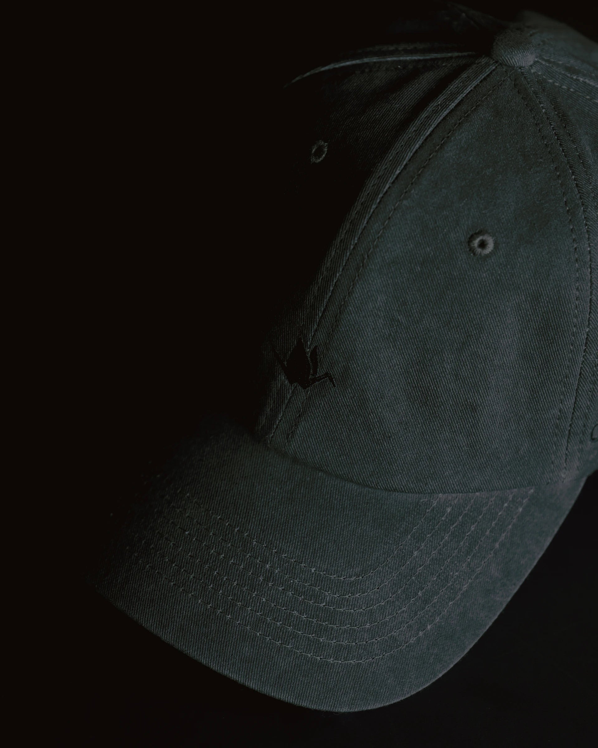 washed black signature dad cap close-up showing curved brim and panel construction, soft cotton fabric with subtle crane embroidery, minimal product detail shot on dark background